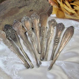 May include: A collection of eight silver-toned metal butter knives or spreaders, each featuring distinct ornate embossed handle designs, including floral, scrollwork, and shield motifs, arranged in a fan shape on a white fabric surface.