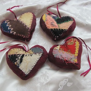 以下が含まれることがあります： 赤、茶色、白の色合いの手作りの布製のハートが4つ。それぞれのハートはパッチワークの布で作られており、異なるパターンをしています。ハートは白い背景に吊り下げられています。