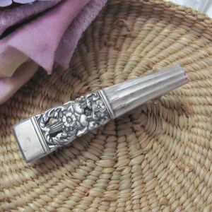 Puede incluir: Un abrecartas antiguo plateado con un diseño floral en el mango y una hoja acanalada. El abrecartas está sobre una cesta tejida, con una tela morada en el fondo.