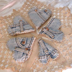 以下が含まれることがあります： ボタンとポケットのディテールが施された、ハート型のデニムオーナメントが5つ、レースのドイリーの上に配置されています。デニムは薄い青色で、ステッチと真鍮色のボタンが見えます。ドイリーは薄いベージュ色です。