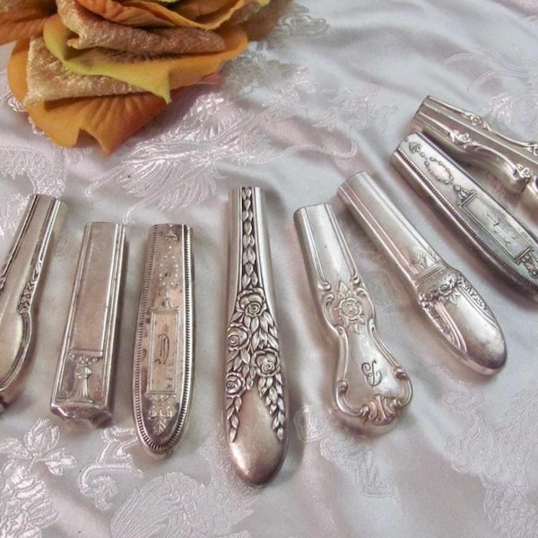 May include: A collection of seven silver-toned metal knife handles arranged in a row. Each handle features distinct embossed patterns, including floral motifs, scrollwork, and geometric designs, with varying shapes and decorative engravings.