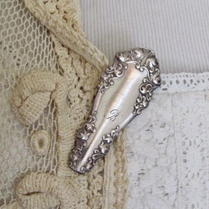 Puede incluir: Un broche de plata, adornado, en forma de cuchara con una letra "B" grabada sobre un fondo de encaje blanco.
