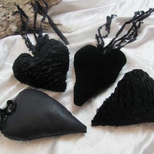 以下が含まれることがあります： フリルのある縁取りが施された黒いベルベットのハート型のクッションが4つ。クッションは白い布の表面に置かれています。