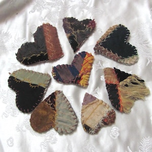 Puede incluir: Una colección de adornos hechos a mano en forma de corazón con patchwork. Cada corazón presenta una combinación única de telas en varios colores, incluyendo negro, marrón y beige, con costuras visibles. Los corazones están dispuestos sobre una superficie blanca texturizada.