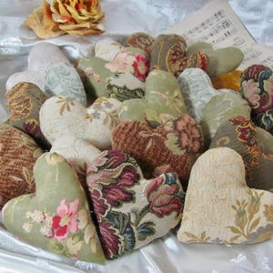 May include: A collection of small, stuffed heart-shaped ornaments crafted from various woven fabrics. The hearts feature diverse patterns including floral motifs, damask prints, and solid textures in shades of sage green, cream, burgundy, and brown.