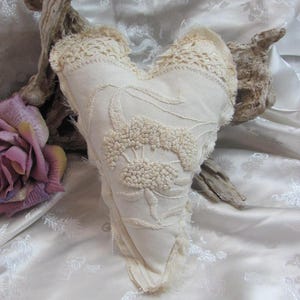 May include: A white heart-shaped pillow with a crocheted edge and intricate embroidered details. The pillow is made of a soft, textured fabric and has a vintage aesthetic.