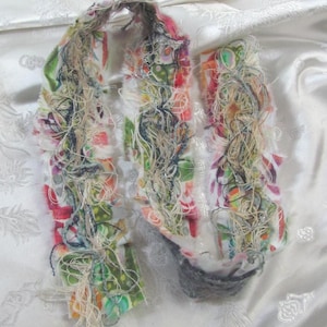 May include: A long, narrow textile strip featuring a white base with a multicoloured floral and abstract pattern. The edges are frayed, and the surface is heavily embellished with a dense, tangled layer of loose, metallic, and neutral-toned threads.
