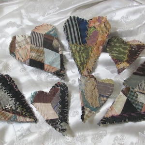 Puede incluir: Una colección de adornos de corazón de tela hechos a mano. Cada corazón está hecho de forma única con varias telas estampadas en tonos de marrón, azul, verde y crema. Los corazones están dispuestos sobre una superficie blanca texturizada.