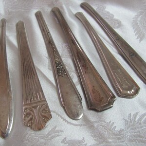 May include: Six silver-plated butter knives with ornate handles. The knives have different designs, including floral, geometric, and a design with the words "The Sheffield American" on the handle.