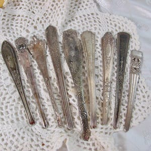 May include: A collection of eight silver-toned metal utensil handles arranged in a fan shape. Each handle features distinct embossed patterns, ranging from floral and scrollwork designs to geometric lines, with varying degrees of metallic patina.