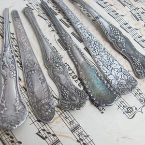 Op de afbeelding: Een assortiment antieke zilveren serveerbestekken met sierlijke bloemen- en druivenontwerpen. De lepels en serveerstukken zijn gerangschikt op een achtergrond van vintage bladmuziek, waarbij hun ingewikkelde details en verouderde patina worden getoond.