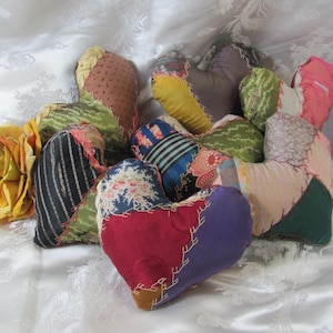 Puede incluir: Una colección de almohadas hechas a mano con forma de corazón y patchwork en varios colores y patrones. Las almohadas están hechas de diferentes telas y cosidas con costuras visibles. Una rosa amarilla se coloca cerca de las almohadas.