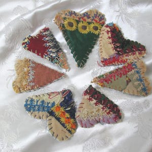 Puede incluir: Colección de adornos de tela hechos a mano en forma de corazón. Cada corazón presenta diseños de patchwork únicos con varios colores, texturas y bordados. Un corazón tiene girasoles. Los corazones están dispuestos en un patrón circular sobre un fondo blanco.