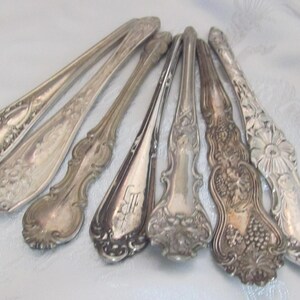 May include: A collection of antique silver spoons with ornate designs. The spoons vary in style, with floral, grape, and monogram details. The spoons are arranged on a white surface, showcasing their intricate patterns and aged patina.