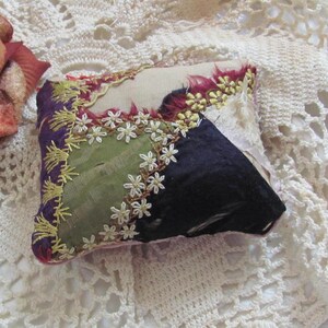 May include: A small, square, patchwork pincushion with a variety of fabrics and embroidery. The pincushion is made of a mix of purple, green, and black fabrics, with white embroidery around the edges. The pincushion is sitting on a white crocheted doily.