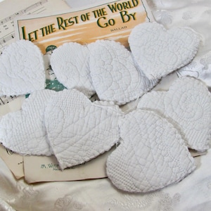 May include: A collection of white heart-shaped fabric ornaments featuring various quilted textures and patterns. Each heart is crafted from white textile material with visible stitching details, arranged in a cluster over vintage sheet music.
