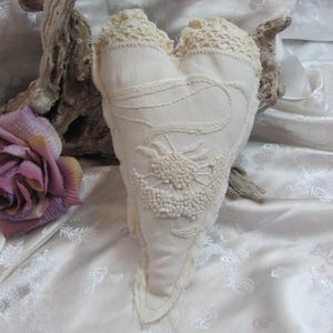 May include: Cream-colored heart-shaped pillow with embroidered floral details and lace trim. The pillow is made of a textured fabric and has a rustic, handmade appearance. A purple rose and driftwood are in the background.