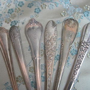 May include: Six silver spoons with ornate designs. The spoons have different patterns, including floral, geometric, and monogrammed designs.