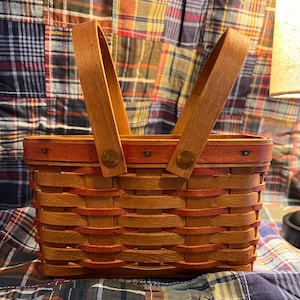 May include: A rectangular woven basket with a wooden handle. The basket is made of light brown and reddish-brown woven strips. The handle is made of light brown wood and is attached to the basket with metal rivets. The basket is sitting on a plaid fabric.