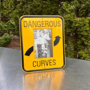 May include: A yellow metal picture frame with the words "DANGEROUS CURVES" in black. The frame has a black border and a black and white photo inside. A black arrow points to the right.