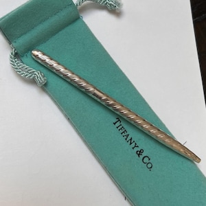 May include: A silver pen with a textured, diagonal pattern rests inside a teal drawstring pouch. The pouch has the words "TIFFANY & Co." printed on it. The pen is a sleek, elegant design.