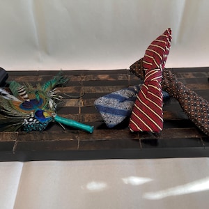 May include: A decorative tray with a black finish, holding several neckties and a feather boutonniere. The ties feature patterns in blue, red, and brown. The boutonniere has green, blue, and yellow feathers.