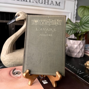 May include: A vintage hardcover book titled L'AVARE by Molière with a dark olive green cloth cover. The front cover features gold embossed heraldic crests at the top, followed by the title and author name in a clean, centered serif font.