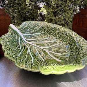 May include: A large, green ceramic serving dish shaped like a cabbage leaf. The dish has detailed veining and a glossy finish. The edges are scalloped, and the overall design is reminiscent of a natural, organic form.