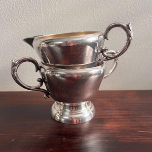 May include: A silver creamer and sugar bowl set. The creamer is stacked on top of the sugar bowl. Both pieces have ornate handles and a shiny finish. The sugar bowl has a pedestal base.
