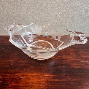 May include: A clear glass serving dish with a tulip design. The dish has a unique shape with handles and an interior compartment. The tulip design is etched into the glass, adding a decorative element to the piece.