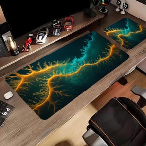 May include: A desk setup with a large rectangular mousepad and a smaller matching pad. The pads have a dark teal background with an abstract design in gold and teal. The wooden desk has toy cars on a shelf. The mousepad is approximately 80 cm long.