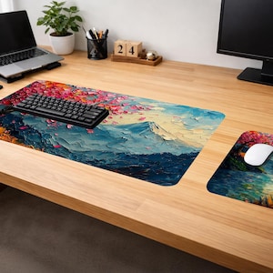 May include: A desk setup with a large rectangular desk mat and a smaller mousepad, both displaying a colourful mountain landscape. The artwork features blue, white, and orange tones, with a black keyboard and white mouse. The desk is made of light-coloured wood.