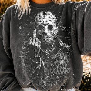 May include: A charcoal grey crewneck sweatshirt featuring a white distressed graphic of a hockey mask-wearing character giving the middle finger. The design includes the text "classy corner club" in a cursive script font on the lower right side.