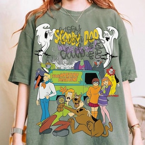 May include: An olive green t-shirt featuring a graphic print of the Scooby-Doo cast, the Mystery Machine van, and ghosts. The text reads Scooby-Doo Where Are You and The Mystery Machine. The design includes characters like Fred, Shaggy, Velma, Daphne, and Scooby-Doo.