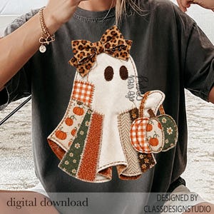 Könnte beinhalten: Ein anthrazitfarbenes T-Shirt mit einer Grafik eines weißen Geistes, der mit einer Schleife im Leopardenmuster verziert ist. Der Geist besteht aus verschiedenen stoffartigen Flicken in Orange, Grün und Weiß und hält zwei kleine Kürbisornamente.