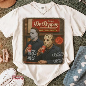May include: A white short-sleeve t-shirt featuring a graphic print of Michael Myers and Jason Voorhees holding red Dr. Pepper cans. Above them is a red sign that reads Drink Dr. Pepper GOOD FOR HALLOWEEN. The shirt has a crew neckline.