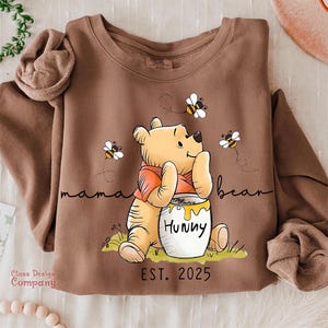 Może przedstawiać: Brązowa bluza z nadrukiem kresk&oacute;wkowego Winnie the Pooh trzymającego słoik miodu z napisem "Hunny". Tekst "mama bear" jest napisany nad misiem, a "EST. 2025" pod misiem.