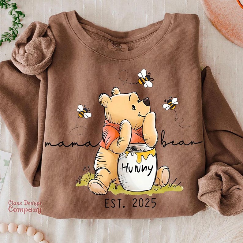 Może przedstawiać: Brązowa bluza z nadrukiem kresk&oacute;wkowego Winnie the Pooh trzymającego słoik miodu z napisem "Hunny". Tekst "mama bear" jest napisany nad misiem, a "EST. 2025" pod misiem.