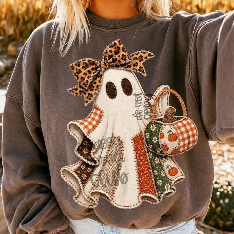 May include: A charcoal gray crewneck sweatshirt featuring a central graphic of a white ghost wearing a leopard print bow. The ghost is holding a pumpkin-shaped basket with orange gingham and floral patterns, and the ghost's body is composed of various fabric-like patches.