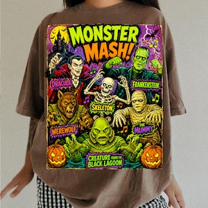 May include: A brown t-shirt featuring a colorful graphic print with the text MONSTER MASH! and illustrations of Dracula, Frankenstein, a skeleton, a werewolf, the mummy, and the creature from the black lagoon, along with two jack-o'-lanterns.