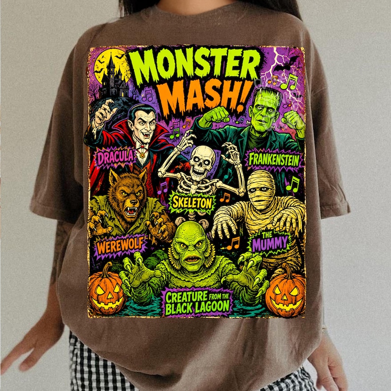 May include: A brown t-shirt featuring a colorful graphic print with the text MONSTER MASH! and illustrations of Dracula, Frankenstein, a skeleton, a werewolf, the mummy, and the creature from the black lagoon, along with two jack-o'-lanterns.