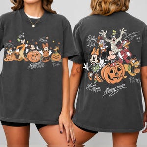 K&ouml;nnte beinhalten: Ein anthrazitfarbenes T-Shirt mit Disney-Figuren in Halloween-Kost&uuml;men. Die Vorderseite zeigt Goofy, Mickey, Minnie und Pluto mit K&uuml;rbissen. Die R&uuml;ckseite zeigt Daisy, Goofy, Donald, Minnie, Mickey und Pluto mit Unterschriften und Text.