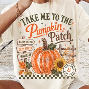 Pode incluir: Uma t-shirt de cor creme com um gr&aacute;fico que inclui o texto "TAKE ME TO THE PUMPKIN PATCH". O design apresenta uma ab&oacute;bora laranja grande, um girassol, folhas de outono, uma placa de madeira com "FARM FRESH", "DAILY PICKED", "PUMPKINS" e uma cerca.
