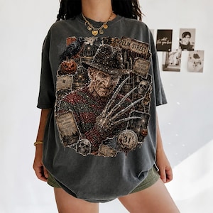 May include: A charcoal gray oversized t-shirt featuring a graphic print of a character with a fedora, a red and green striped sweater, and bladed fingers. The design includes text reading Elm Street, Welcome to your nightmare, and 31.