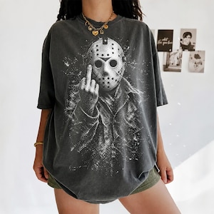 Puede incluir: Una camiseta gris carb&oacute;n de corte oversize con un estampado gr&aacute;fico blanco desgastado de una figura con m&aacute;scara de hockey haciendo un gesto de dedo medio. La prenda tiene mangas cortas, cuello redondo y un acabado de estilo vintage.