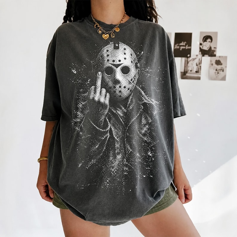 Puede incluir: Una camiseta gris carb&oacute;n de corte oversize con un estampado gr&aacute;fico blanco desgastado de una figura con m&aacute;scara de hockey haciendo un gesto de dedo medio. La prenda tiene mangas cortas, cuello redondo y un acabado de estilo vintage.