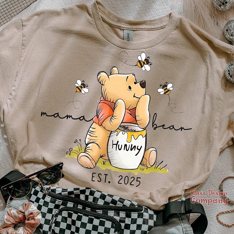 Może przedstawiać: Beżowa koszulka z nadrukiem kresk&oacute;wkowego obrazka Winnie the Pooh trzymającego słoik miodu. Na koszulce nadrukowany jest tekst "mama bear" i "Hunny". Pod obrazkiem nadrukowany jest tekst "EST. 2025".