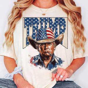 May include: A white t-shirt with a vintage-style graphic of a man wearing a cowboy hat with the American flag on it. The graphic also includes the word "TRUMP" in a star-shaped design.