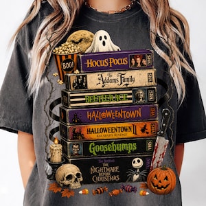 May include: A charcoal gray t-shirt featuring a graphic print of a stack of VHS tapes titled Hocus Pocus, The Addams Family, Beetlejuice, Halloweentown, Halloweentown II, Goosebumps, and The Nightmare Before Christmas, with a skull, candle, and pumpkin.