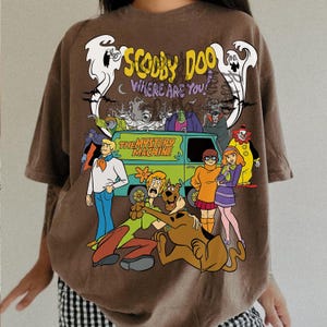 May include: A brown t-shirt featuring a graphic print of the Scooby-Doo cast, the Mystery Machine van, and ghosts. The text reads Scooby Doo Where Are You and The Mystery Machine. The design includes various characters like Shaggy, Velma, and Daphne.